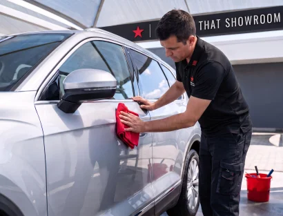 A man in a black uniform with a red star logo carefully buffs the side door of a silver SUV using a red cloth, under an outdoor detailing canopy.