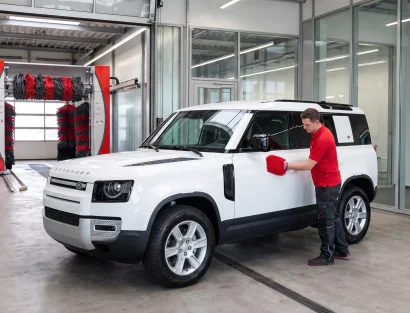 A worker in a red shirt hand-washes a white Land Rover Defender with a red mitt, parked beside an automatic car wash tunnel with red and black brushes.