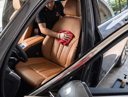 A detailer in a black branded polo shirt uses a red microfiber cloth to clean tan leather seats through the open driver's door of a black car.