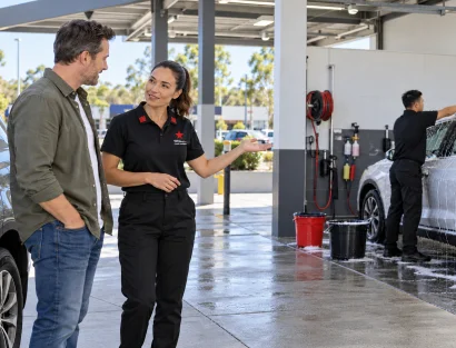 A female car wash attendant in a black branded uniform gestures toward the wash bays while talking with a male customer standing beside his SUV.