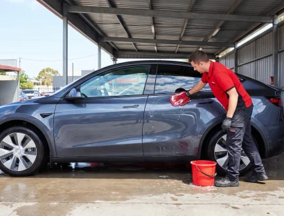 A man in a red uniform hand-washes a gray Tesla Model Y under an open-air canopy, with another worker cleaning a second car in the background.